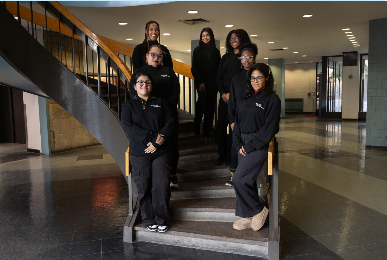 A professional group portrait of seven diverse young women standing on the steps of a wide, industrial-style staircase. The group is arranged at different levels of the stairs, creating a tiered composition. They are dressed in cohesive all-black attire, including quarter-zip sweatshirts and trousers. The setting is a brightly lit, spacious lobby with polished floors and large windows.