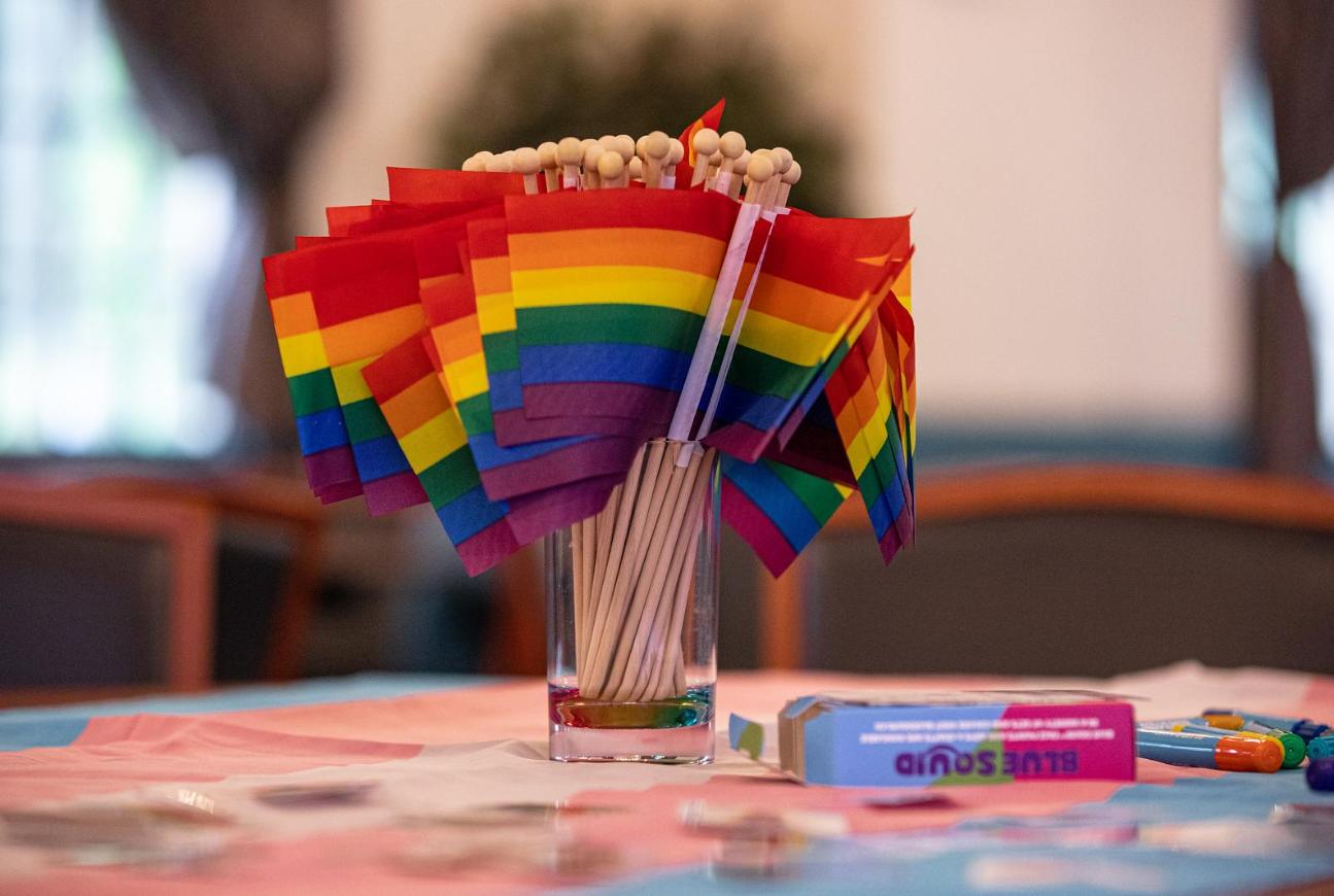 Small rainbow Pride flags displayed in a clear glass on a table covered with a blue, pink, and white Transgender Pride flag.