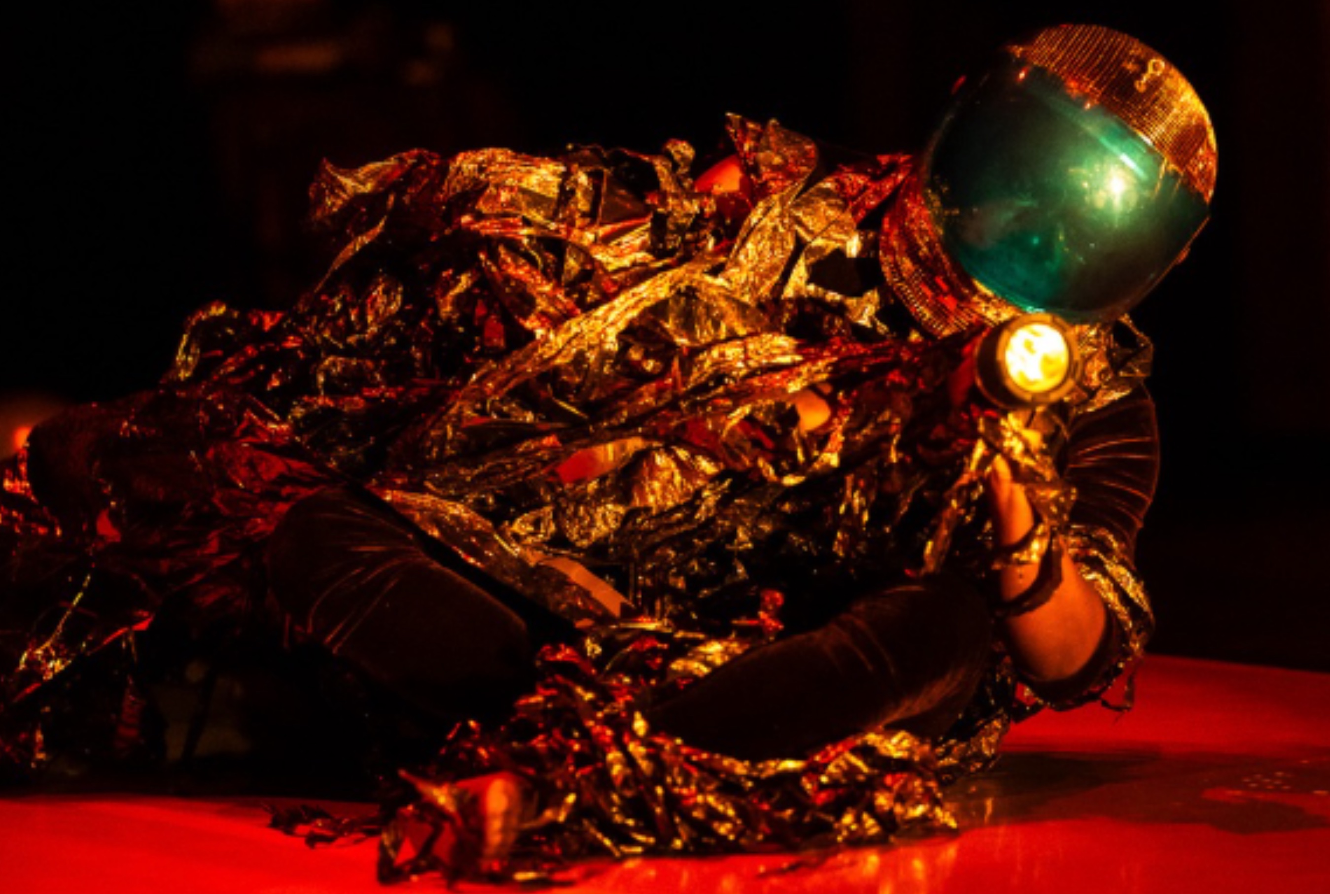 A performer lays on a red floor pointing a flash light towards the audience. The performer is wearing an astronaut-like helmet and a gold mylar costume.