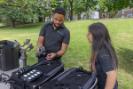 Two Multimedia crew members reviewing media equipment and a camera outdoors on a grassy lawn.