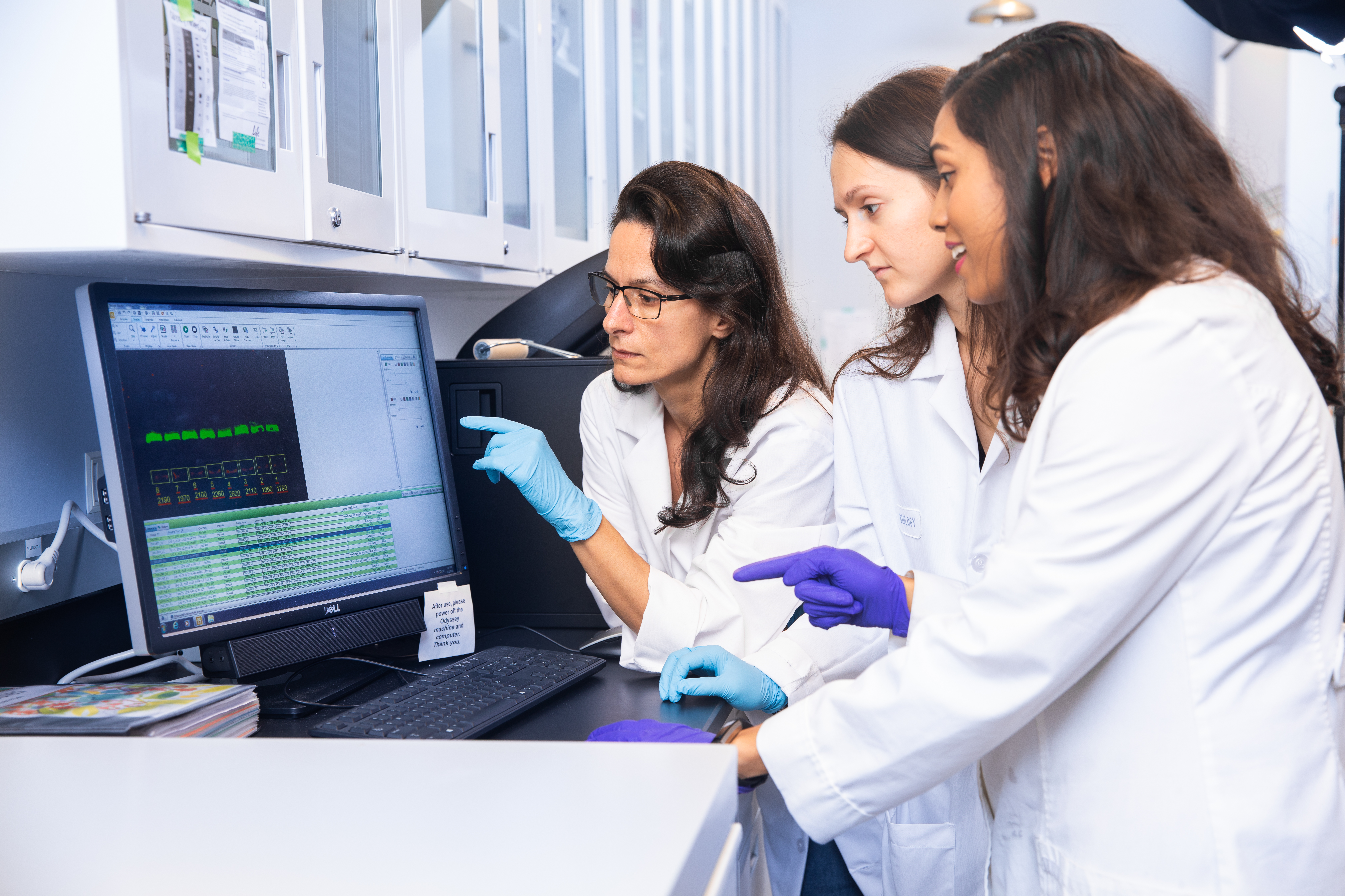 Two female students and female professor wearing white coats in a laboratory looking at a computer monitor.