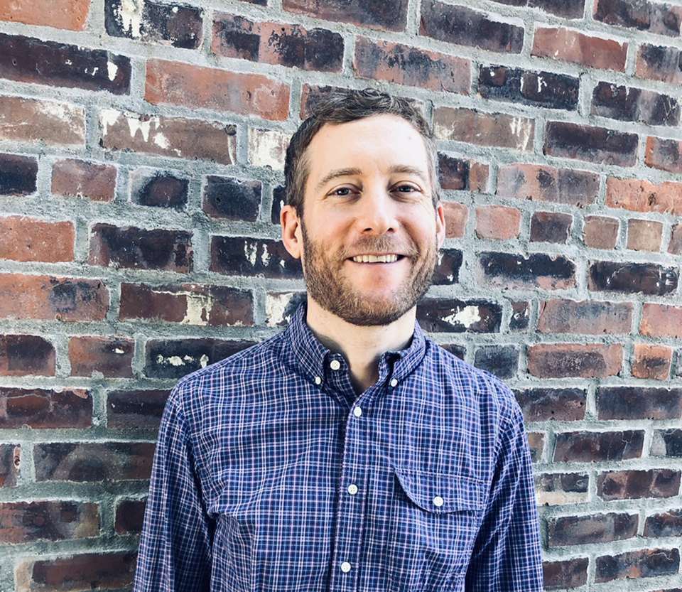 Smiling, bearded man in a blue plaid shirt standing in front of a brick wall.