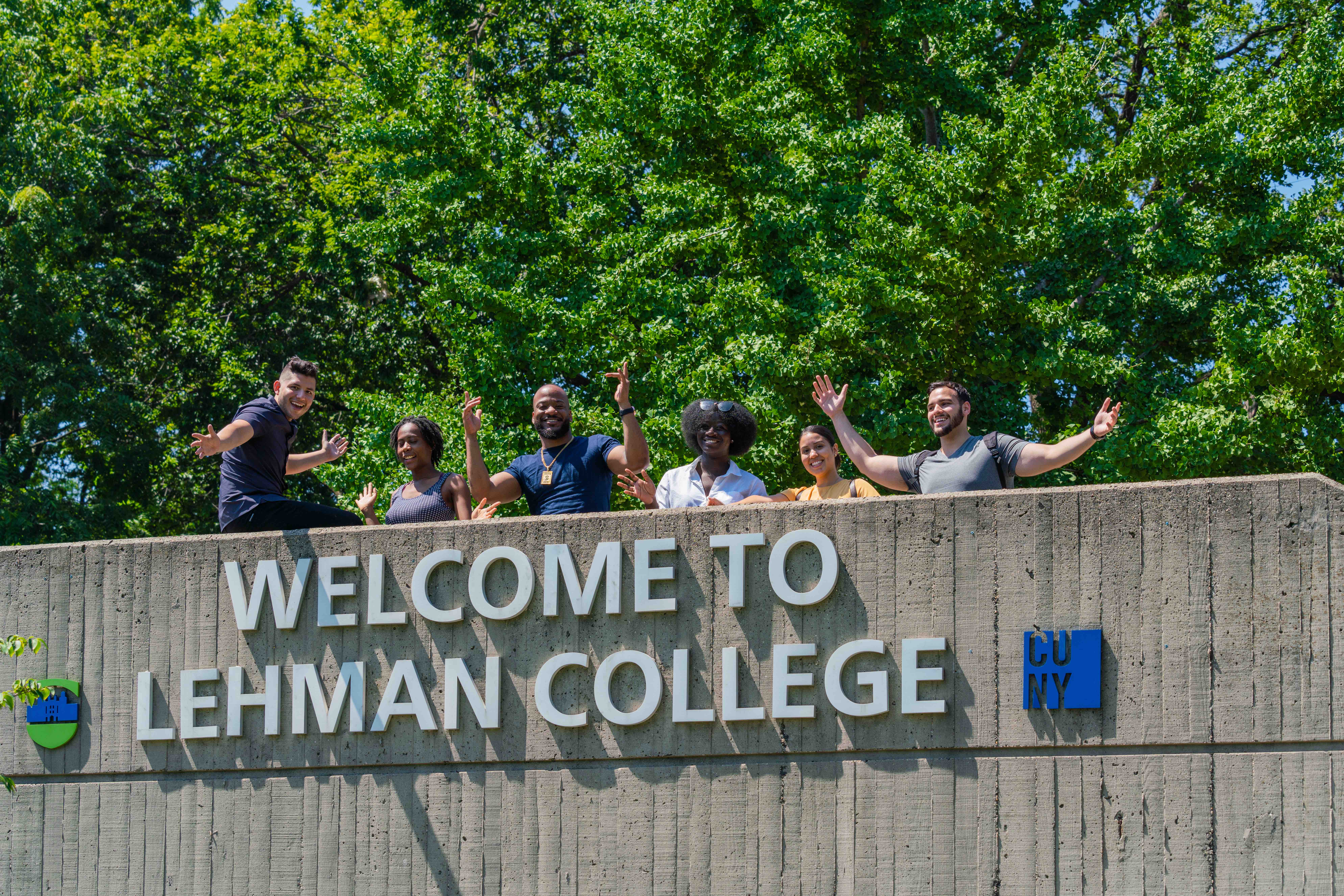 Students standing behind the Welcome to Lehman College sign and waving.