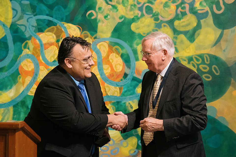 Two men in dark suits shaking hands in front of a green and yellow abstract painting.