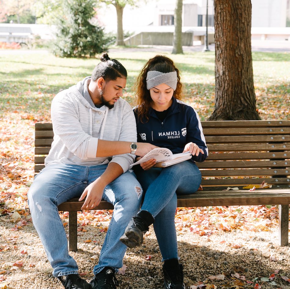 A man and woman sitting on a wooden bench under a tree. He is pointing to a page in a book on her lap.