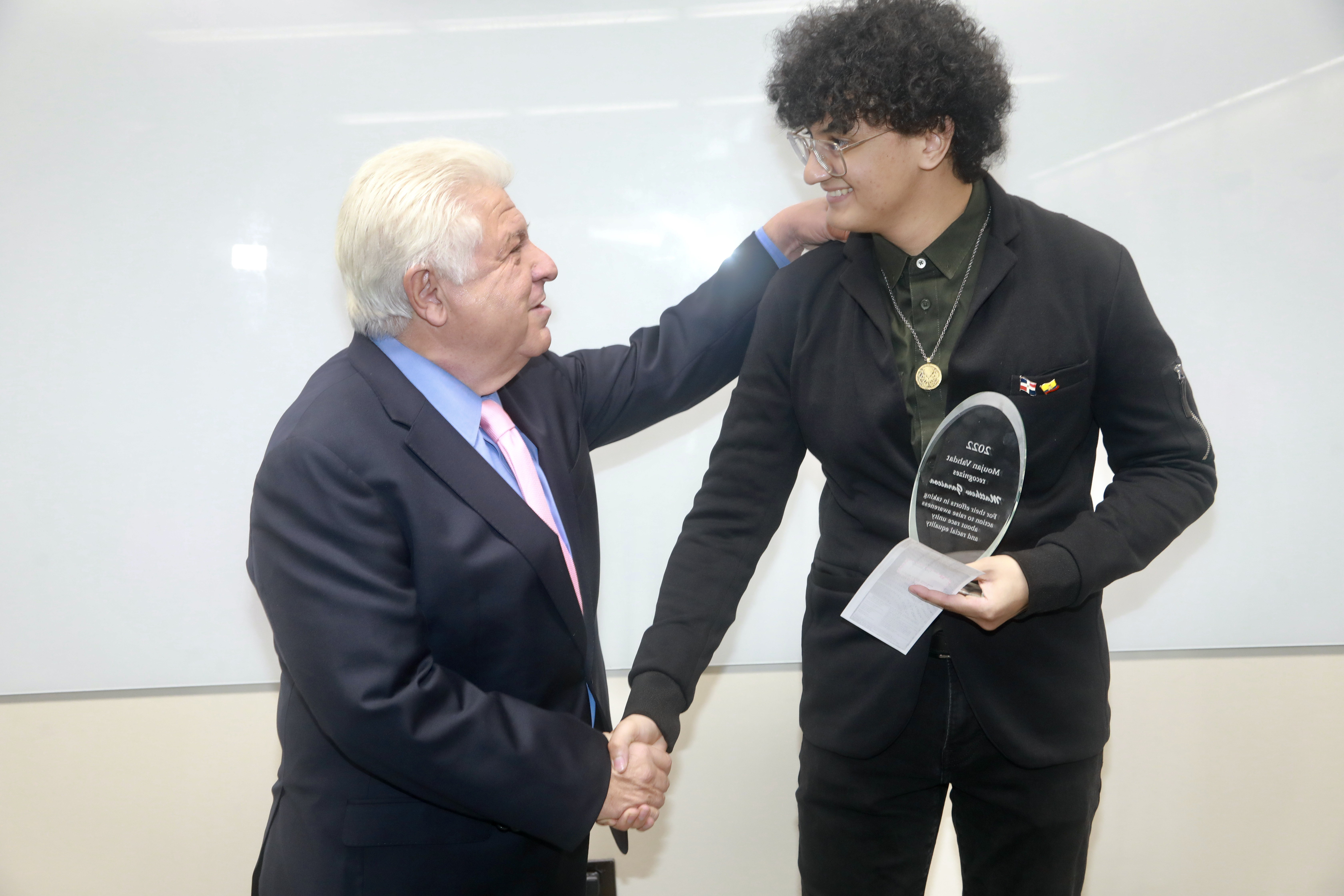 An older man with white hair wearing a suit presents an award to a tall, young man with curly hair.