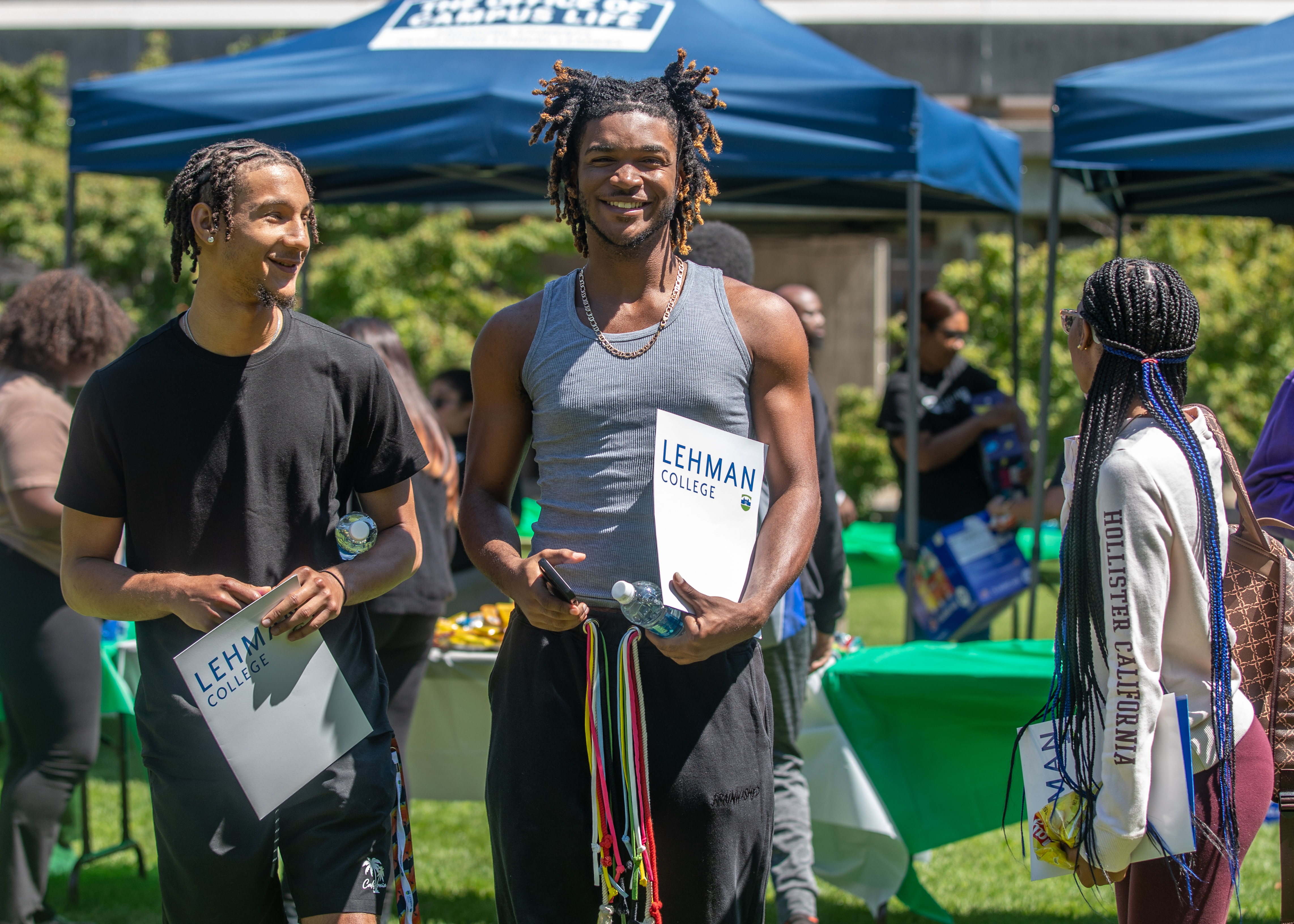 Two men of color standing and smiling at an outdoor event. Both are holding Lehman College folders.