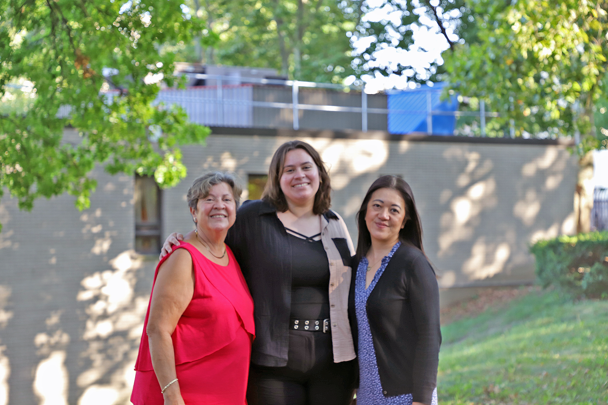 Three women pose for the camera outside with buildings and trees in the background.