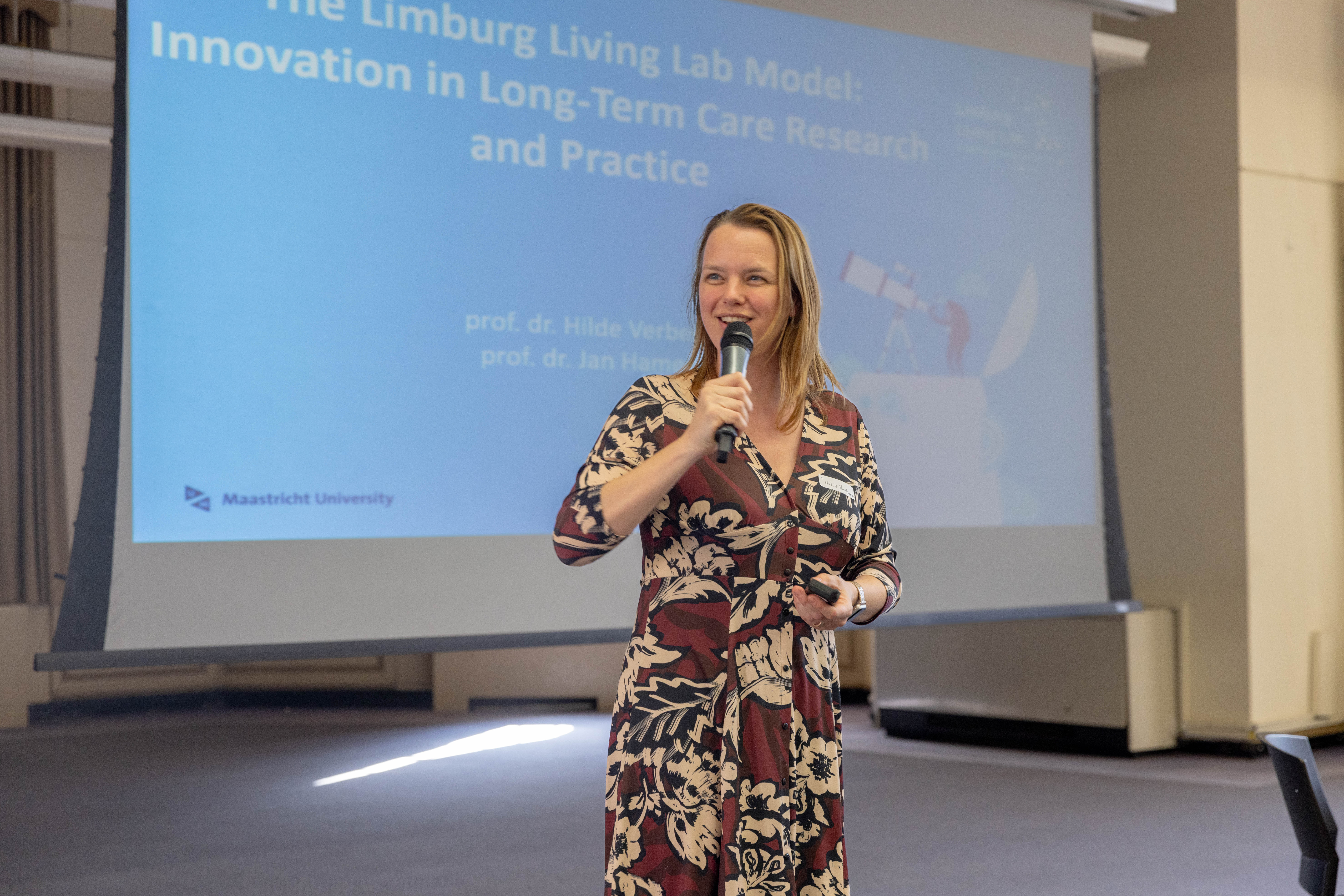 A woman lectures in front of a large screen displaying her talk's title,  The Limburg Living Lab Model: Innovation in Long-Term Care Research and Practice.