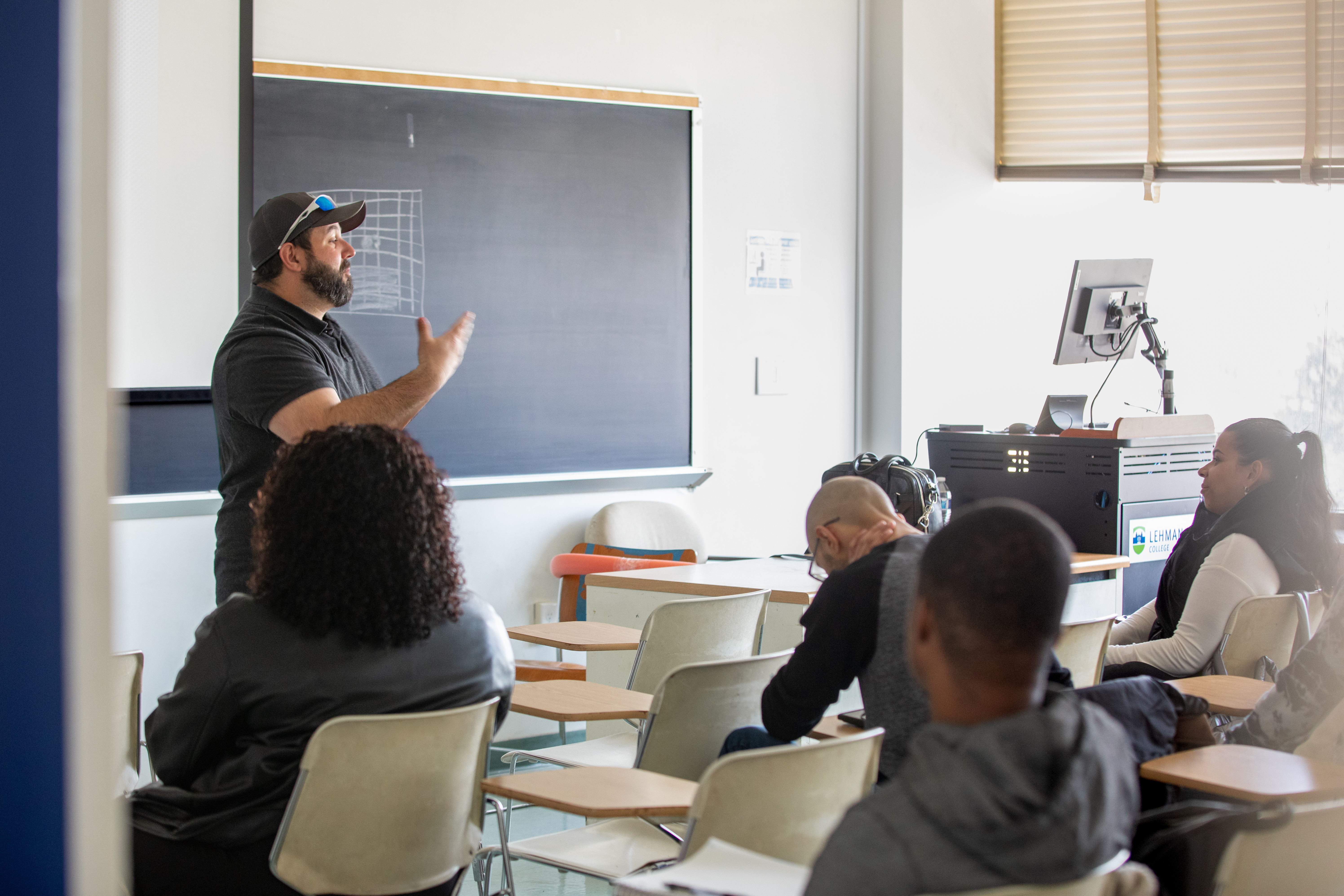 A professor standing and speaking in front of a group of students sitting in a classroom.