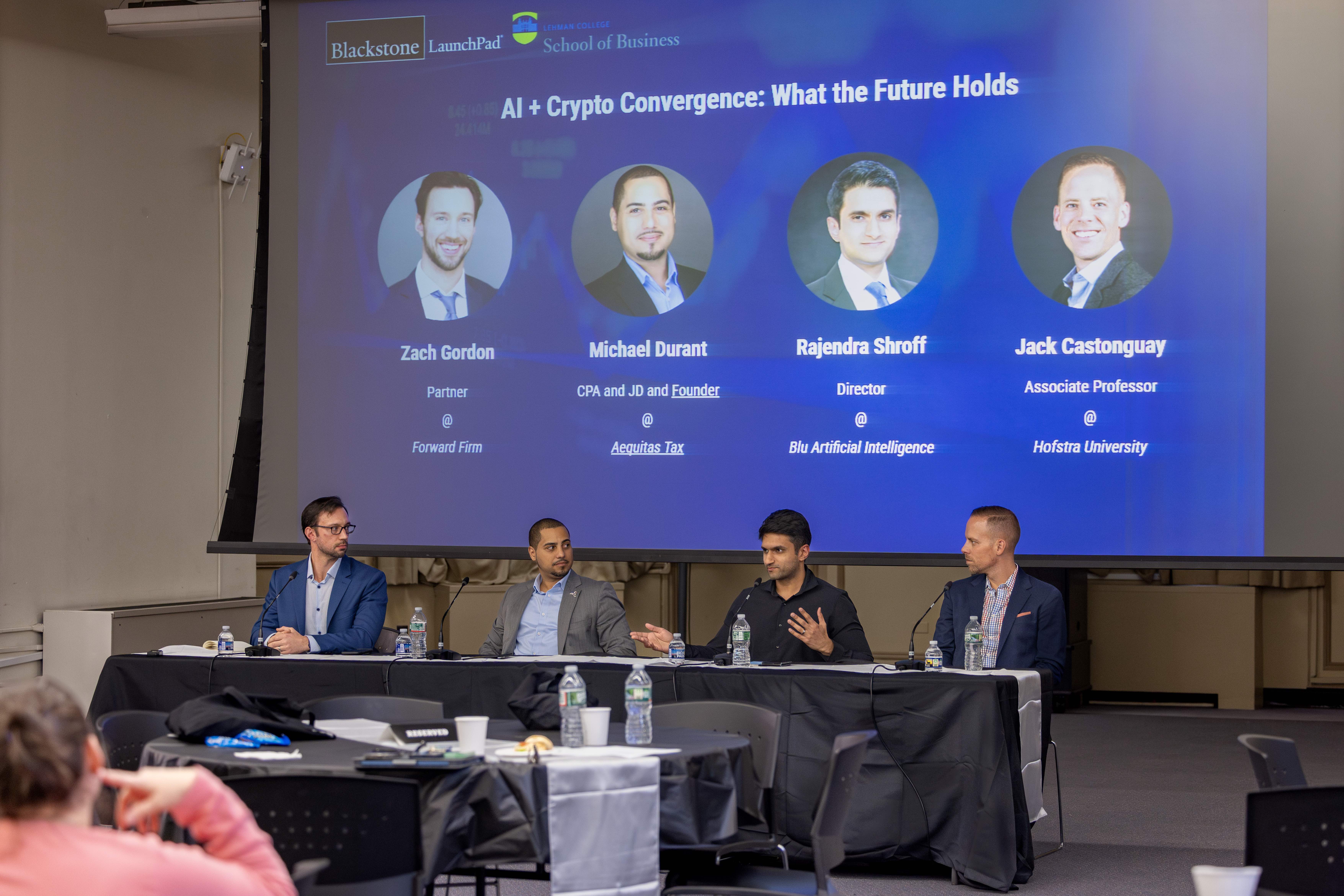 Four men sitting at a table in a panel discussion. A blue backdrop includes their photos and names.