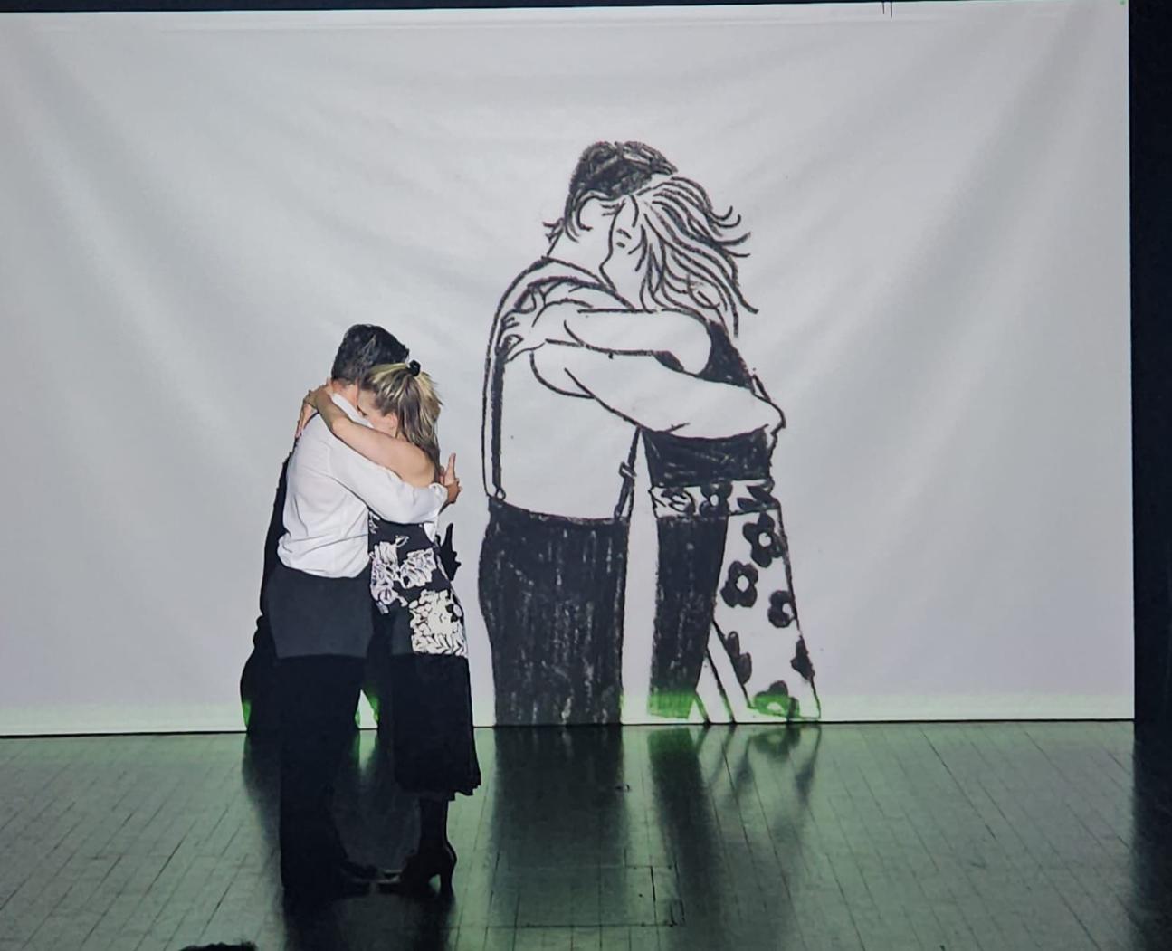 Couple stands closely embracing on a stage in front of a large projected illustration of two people hugging. The live figures mirror the sketched figures behind them, blending reality with the drawn image