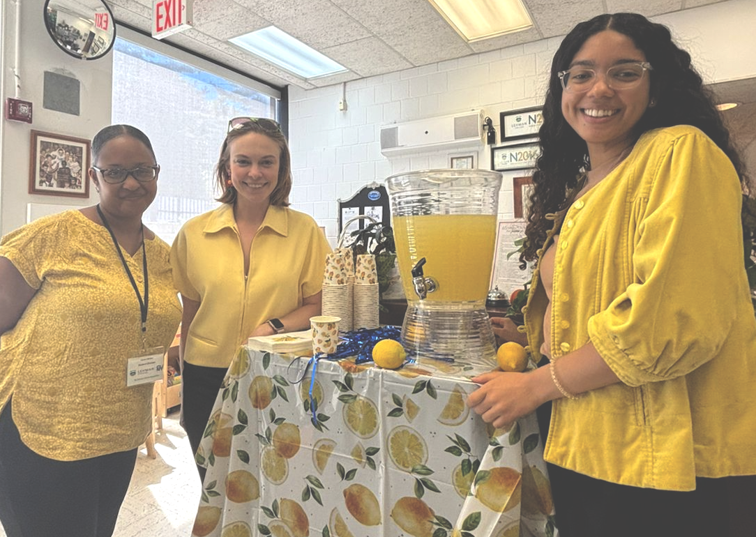 Caron Hinton, Devon Perry, and Ruth Perez Mercado, all in yellow tops, stand next to a table with a dispenser of lemonade.