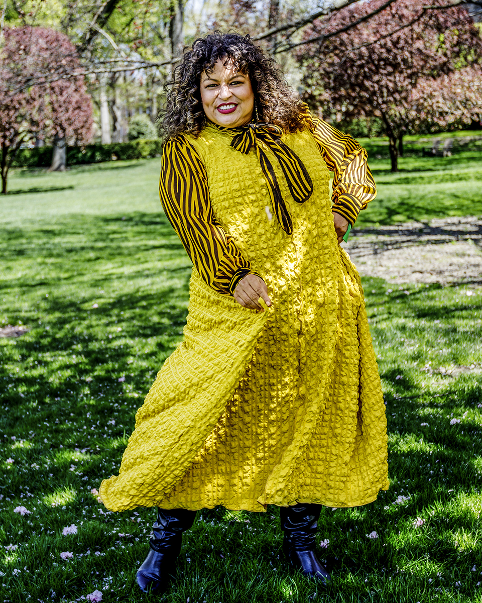 Artistic portrait of a woman in a long yellow dress outside on grass with blooming trees in the background.