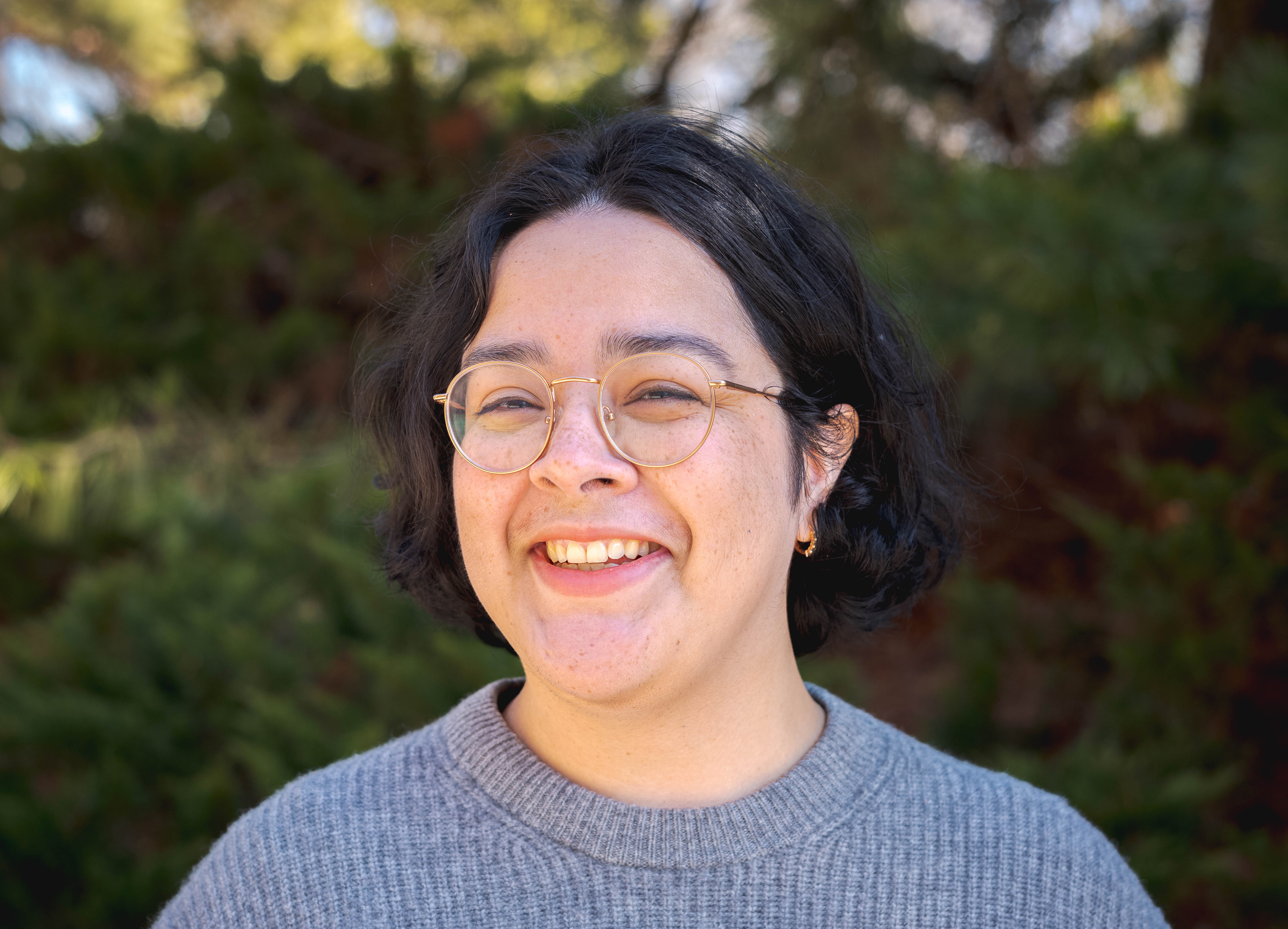 FEMALE WITH SHORT HAIR, SMILING, WEARING A BLUE SWEATER, GOLD WIRE-RIMMED GLASSES, AND SMALL GOLD HOOP EARINGS. STANDING INFRONT OF GREEN BRUSH