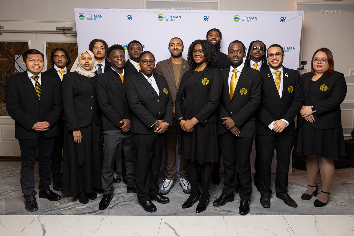 A professional group portrait of students and staff from the Lehman College Urban Male Leadership Program, wearing black blazers with gold crests and gold ties, posing in front of a Lehman College backdrop.